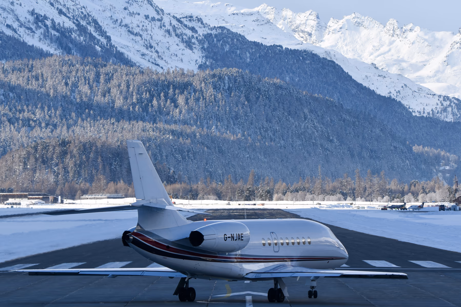 A Falcon private jet at rest on a plowed alpine apron, snow-capped mountains rising behind